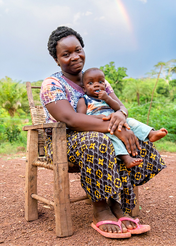 An eco-guard sits in a chair with her young child. In the background is lush greenery and a rainbow.